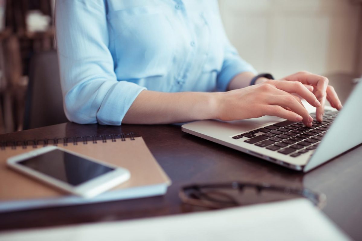 A person photographed from the neck down, with their hands typing on a laptop and a notepad and phone laid on the desk