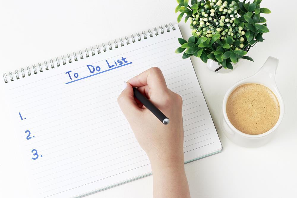 Person writing a to do list with a cup of coffee and a plant on the desk