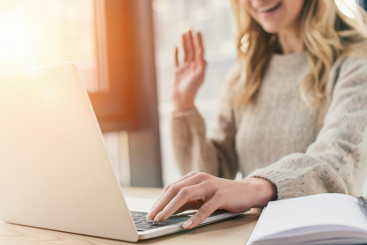 A lady waving at her laptop, with her left hand resting on the keyboard and a notepad and pen on the desk next to her